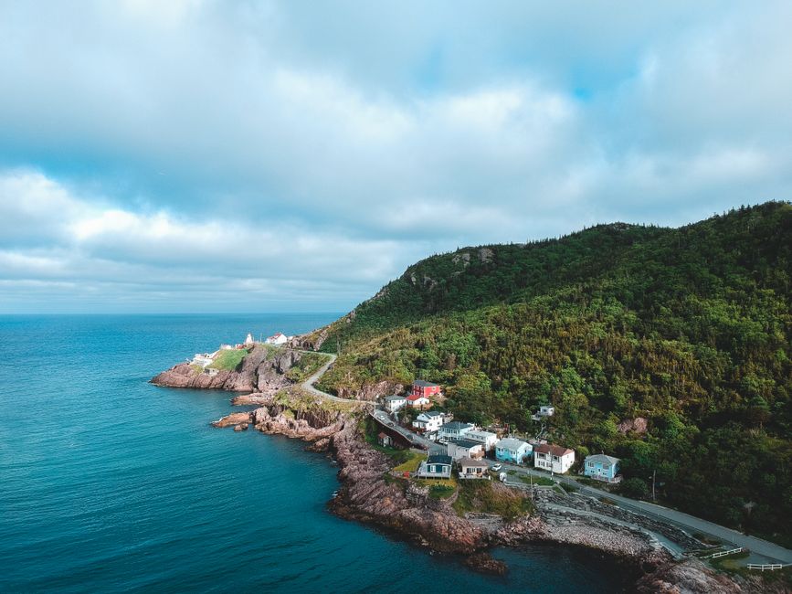 Aerial view of a coastal village with a narrow winding road running alongside the shoreline, leading up a slope densely covered with green trees and foliage. The foreground features rocky cliffs and a small collection of white, grey, and pastel-colored houses with flat or slightly sloped roofs, positioned close to the water's edge. The calm, blue-green sea extends into the distance under a partly cloudy sky with shades of grey and blue. The overall scene depicts a tranquil seaside environment, typical of rural or suburban areas, with no visible signs of waste or rubbish. This landscape provides a serene backdrop contrasted by the man-made structures, and while not directly related to rubbish removal services, it exemplifies a clean coastal setting that could benefit from community or private waste management efforts. The image captures natural beauty and settlement development along the coast, possibly representing a scenic location where rubbish clearance and environmental preservation could be relevant for maintaining the area's appeal.