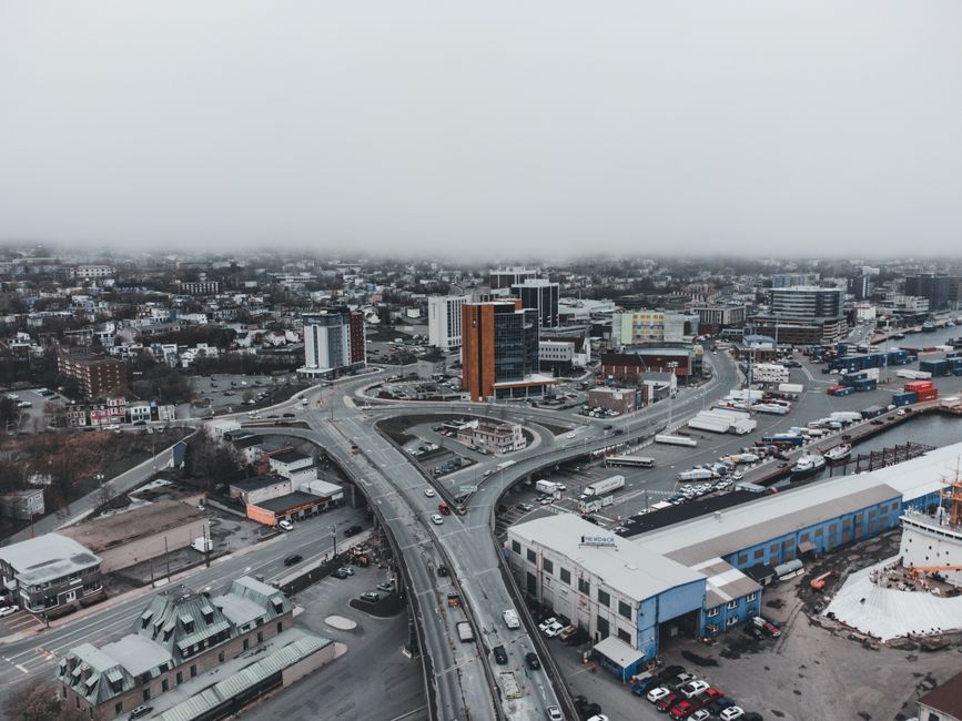 An aerial view of a cityscape shows multiple levels of elevated roads and bridges intersecting in the foreground, with various vehicles traveling along the thoroughfares. Surrounding the roads are commercial and industrial buildings, some with flat roofs and large windows, painted in shades of white, blue, and grey. Behind the infrastructure, a dense cluster of mid-rise office buildings and smaller structures extends into the distance, with the skyline partially obscured by low-hanging grey cloud cover. The environment appears to be a busy urban area with active transportation routes and mixed-use developments. The overall scene captures the functional aspect of city infrastructure, supporting the context of alternative waste handling and urban logistics, aligning with professional rubbish removal services that complement city development and waste management strategies.