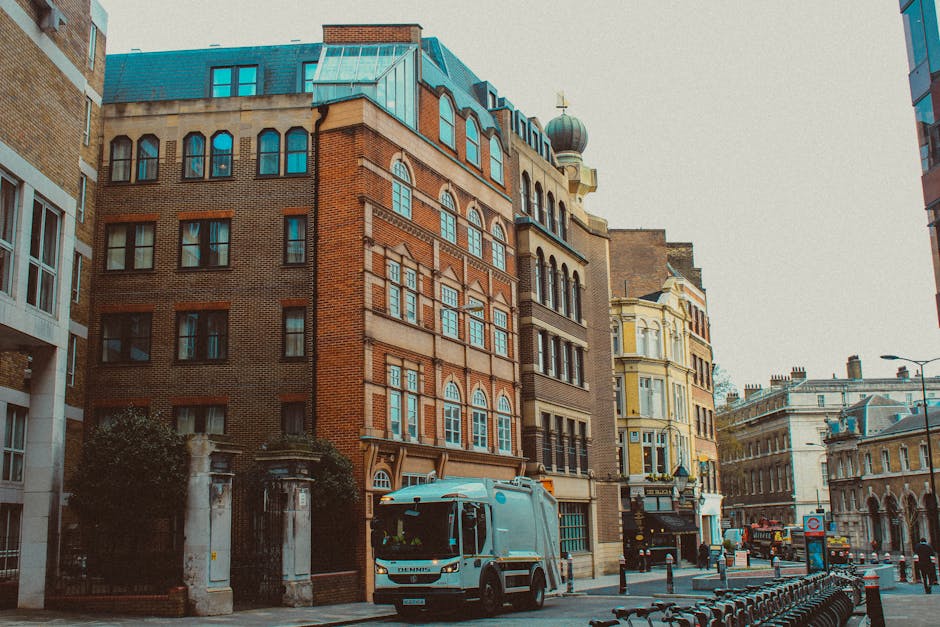 A city street scene showing a white waste collection vehicle parked in front of multi-storey buildings with a combination of brick and ornate facades. The vehicle, positioned in the lower center of the image, is partially shown and appears to be equipped for rubbish collection, consistent with typical private waste handling services. Surrounding structures include buildings with large windows, decorative stonework, and a mix of architectural styles, with some featuring arched windows and detailed cornices. The street is lined with metal bike racks, bollards, and street lamps, with a pavement that appears clean and well-maintained. In the background, additional historic and modern buildings are visible, along with street signage and a few pedestrians, suggesting an urban environment where on-site rubbish collection services are an integral part of waste management. The lighting is natural, indicating daytime conditions, and the scene reflects an active street designated for convenient refuse collection within a busy city area, aligning with private rubbish removal operations.