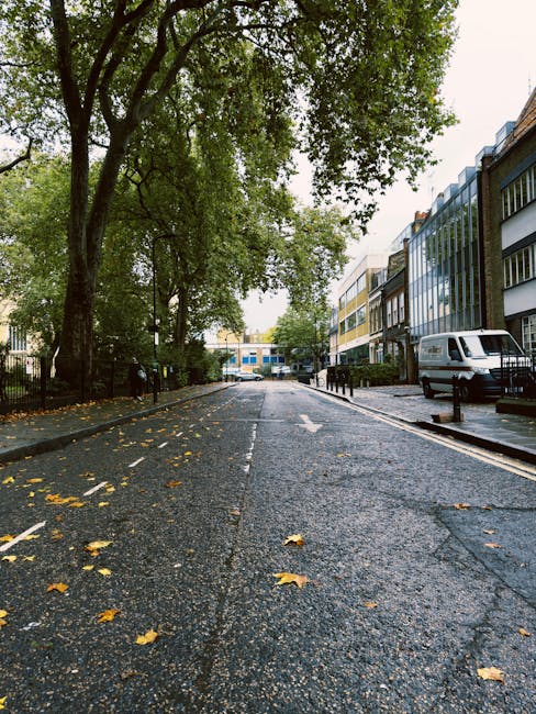 A city street scene showing a white waste collection vehicle parked in front of multi-storey buildings with a combination of brick and ornate facades. The vehicle, positioned in the lower center of the image, is partially shown and appears to be equipped for rubbish collection, consistent with typical private waste handling services. Surrounding structures include buildings with large windows, decorative stonework, and a mix of architectural styles, with some featuring arched windows and detailed cornices. The street is lined with metal bike racks, bollards, and street lamps, with a pavement that appears clean and well-maintained. In the background, additional historic and modern buildings are visible, along with street signage and a few pedestrians, suggesting an urban environment where on-site rubbish collection services are an integral part of waste management. The lighting is natural, indicating daytime conditions, and the scene reflects an active street designated for convenient refuse collection within a busy city area, aligning with private rubbish removal operations.