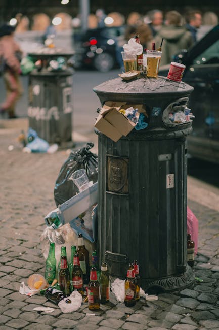 An overflowing public trash bin made of dark metal with a rounded top, placed on a cobblestone street. The bin's lid is open, and various litter such as crumpled paper, fast-food containers, plastic cups, and discarded food wrappers are visible both inside and on top of it. Surrounding the base of the bin, there are several empty glass bottles, some lying on their sides and others upright, with additional paper and plastic waste scattered around. In the background, blurred figures of pedestrians and parked cars suggest a busy urban environment. The scene reflects a typical setting for informal rubbish disposal, illustrating the importance of proper waste management and potentially indicating the need for private waste collection services such as those offered by Rubbish Removal St Johns Wood. The lighting is natural, with soft daylight illuminating the scene, highlighting the textures of the discarded rubbish and the cobbled pavement.
