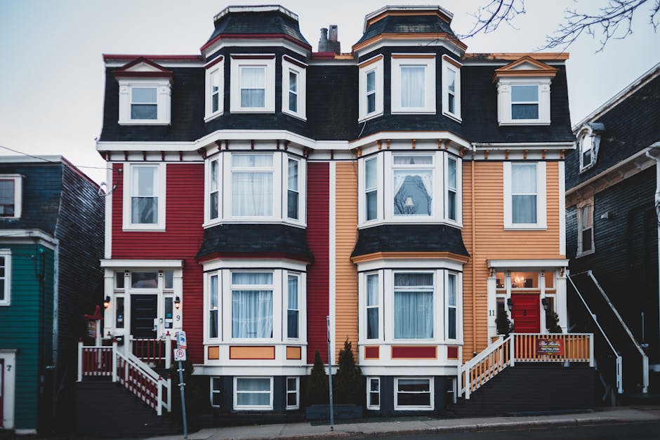 The image depicts a multi-storey Victorian-style residential building with a symmetrical facade, featuring a combination of red and orange wooden cladding accented with white trim. The structure has a distinctive bay window design on both the ground and upper floors, with the bay windows centrally aligned and topped with black, slate-style pitched roofs. The building's exterior includes large, white-framed sash windows, some with sheer curtains visible from the outside. The main entrance, located on the right side, has a red-painted door and a small, accessible staircase with white railings and matching decorative posts. Surrounding the entrance are neatly trimmed small shrubs and potted plants. The building is situated on a paved street with a concrete sidewalk and is flanked by neighboring houses, partly visible on either side. The environment appears to be a residential urban area, with leafless tree branches visible at the top right. The lighting suggests it is either late afternoon or a cloudy day, providing diffuse, natural illumination. This type of building may be associated with private property management, where external rubbish removal services like those from rubbishremovalstjohnswood.co.uk could be involved in on-site clearance and alternative waste handling for property maintenance or renovations.